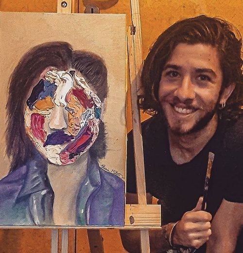 Khevin Mituti, smiling, holding a paintbrush next to one of his oil-painted portraits.